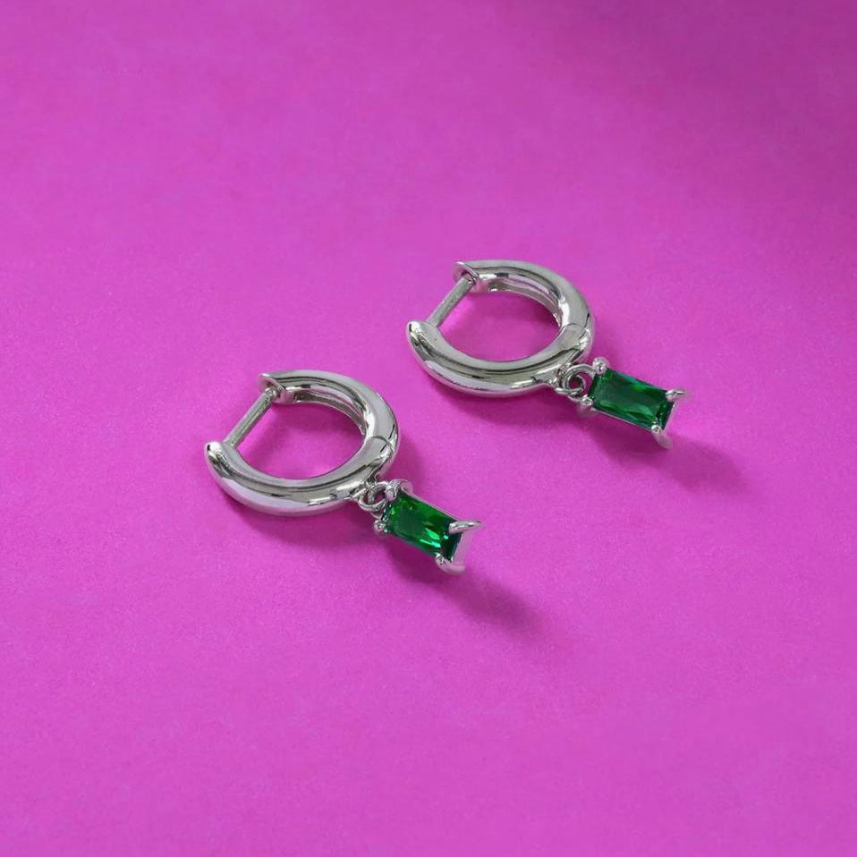 Sterling silver huggie hoop earrings with emerald color stone charms displayed against a bright magenta paper background