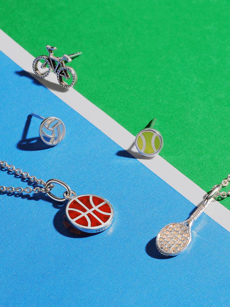 a collection of sterling silver sports jewelry featuring a basketball pendant, a bicycle earring stud, a volleyball stud, a tennis ball and tennis racquet pendant necklace on a green and blue paper background