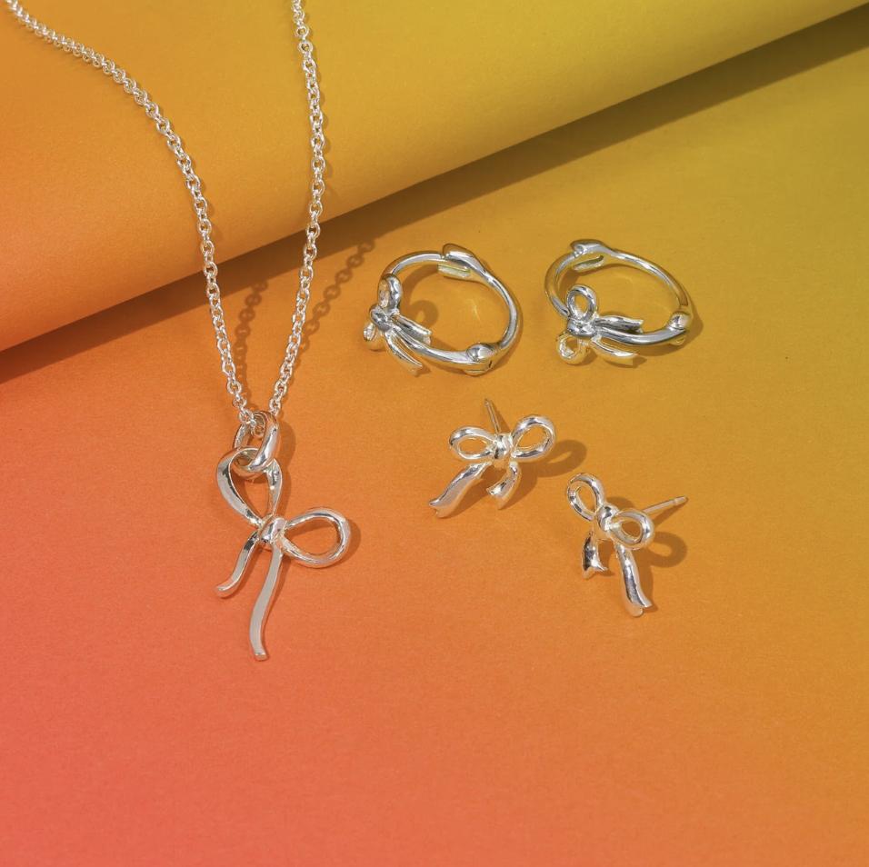 bow tie ribbons collection in sterling silver featuring a necklace, huggies and earring studs displayed against a bright orange and yellow ombre paper background